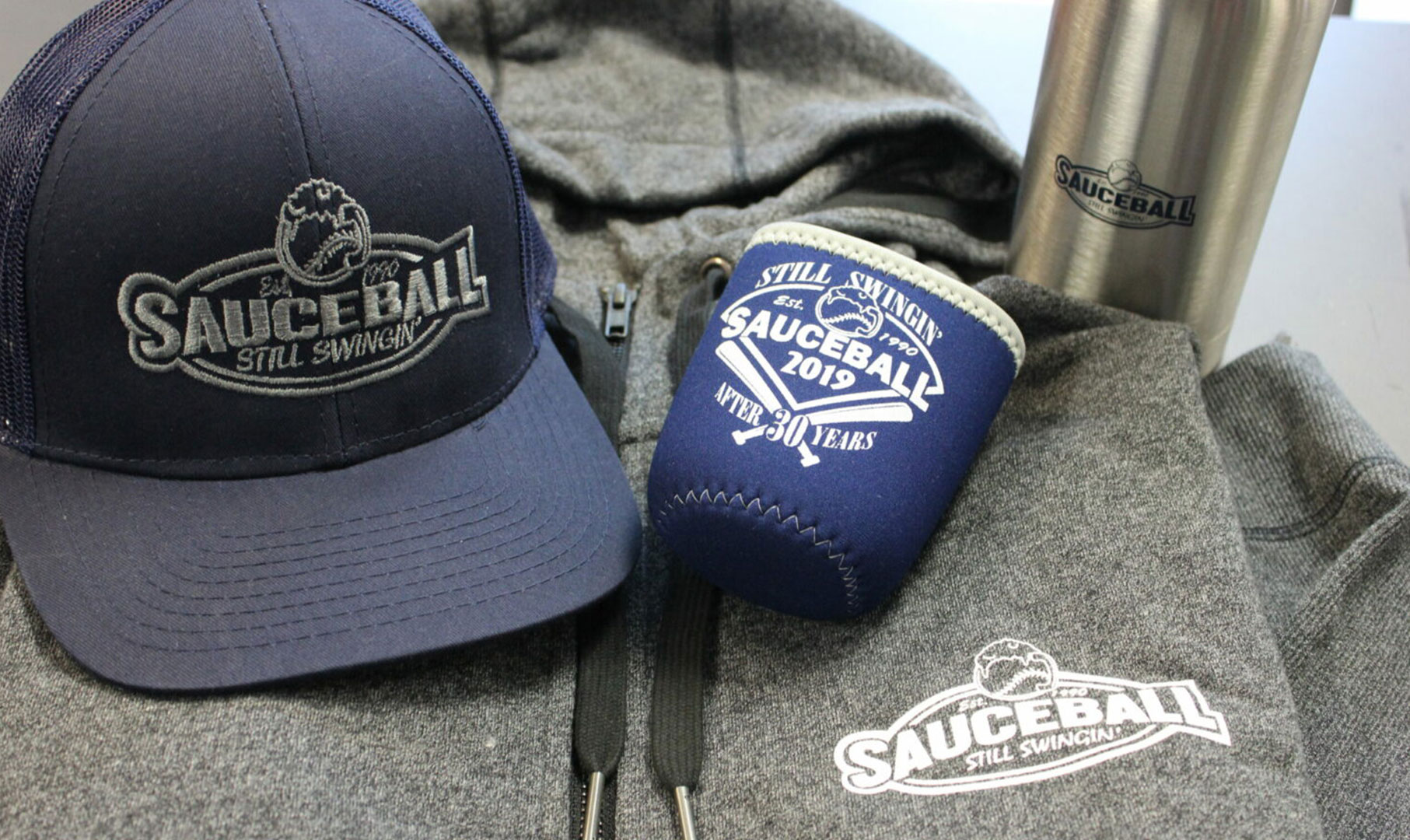 Image of baseball hat, can koozie and hooded sweatshirt laying on a table.