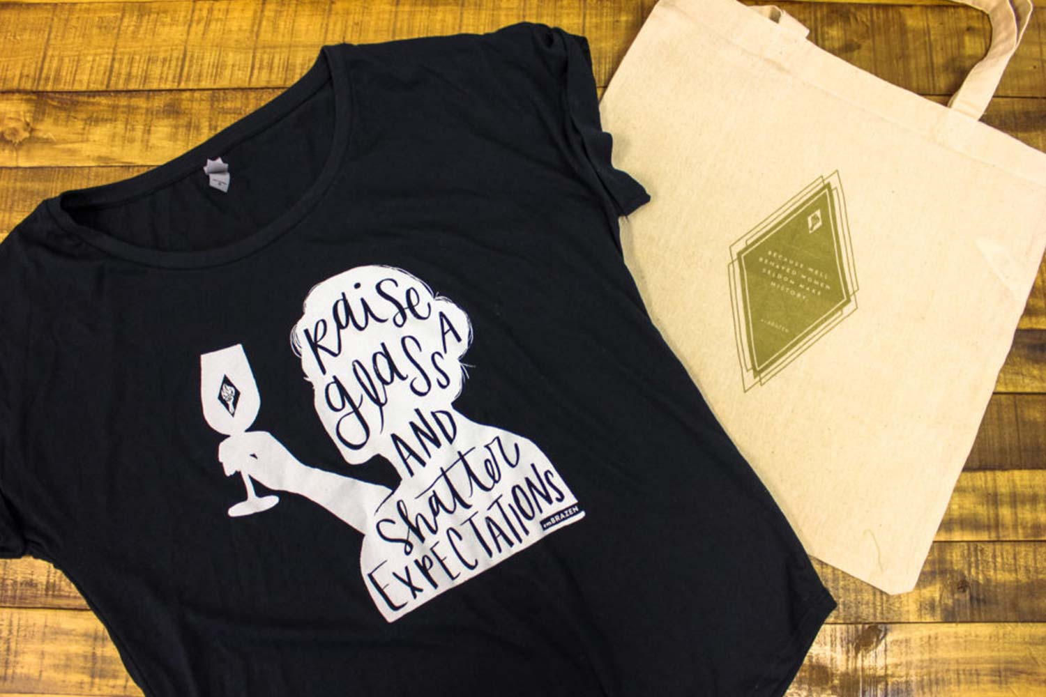 Image of black t-shirt with shadow image of a women and a tote bag with the Embrazen wine logo on it.