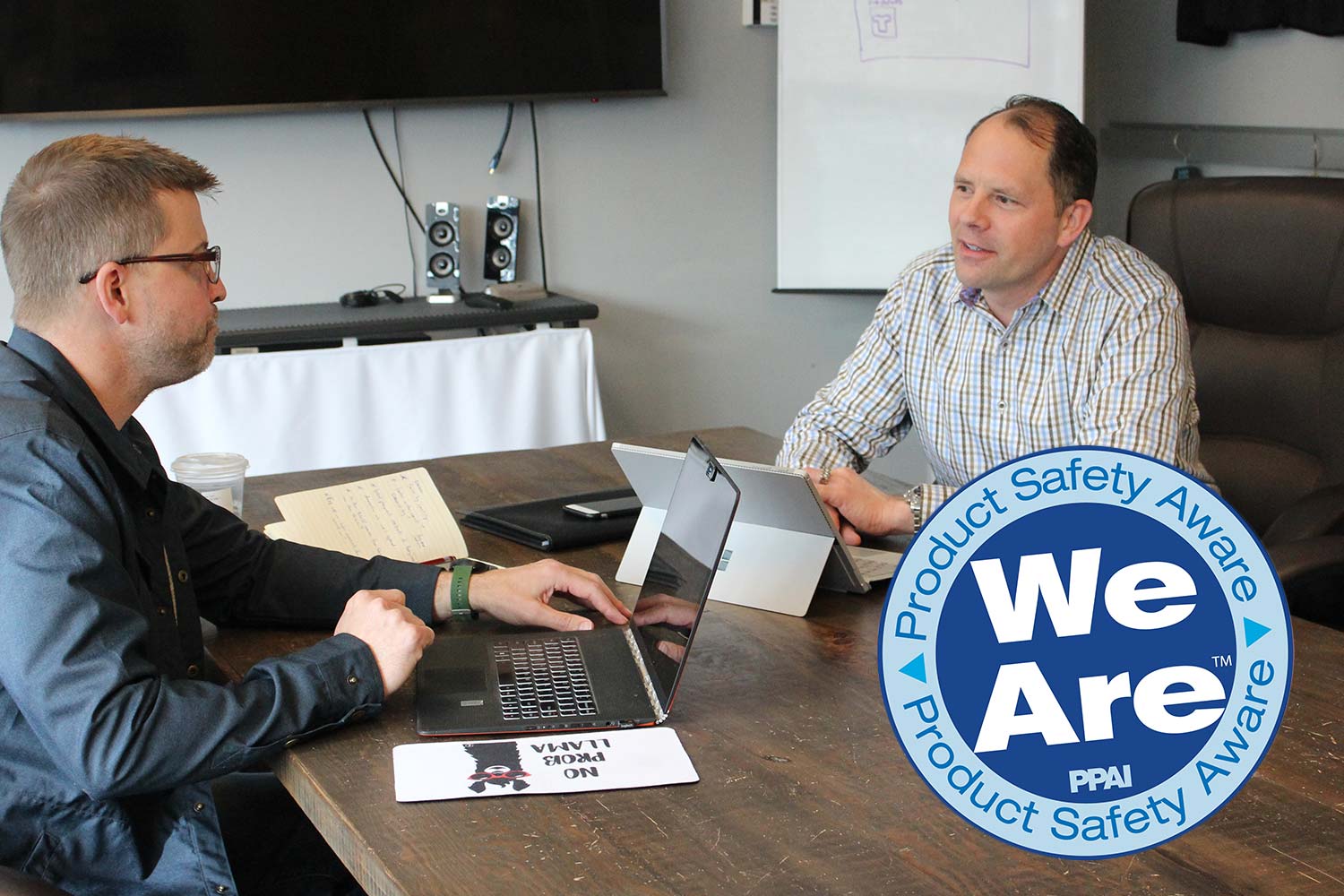 Image to two people meeting in a conference room with PPAI Product Safety Aware logo superimposed on image.